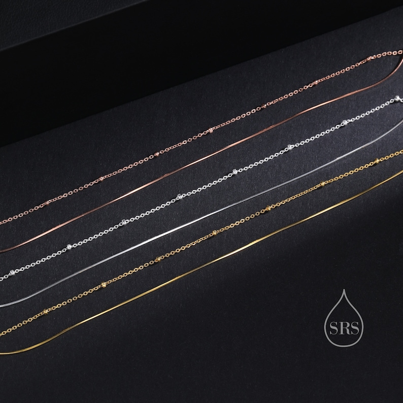 May include: A collection of five delicate necklaces arranged diagonally. The set includes three metal chains with small evenly spaced beads in rose gold, silver, and gold tones, alongside two smooth, thin, solid metal chains in silver and gold.