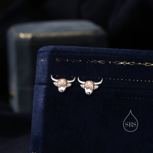 May include: A pair of silver earrings with a Highland Cow design. The earrings are on a blue velvet background.