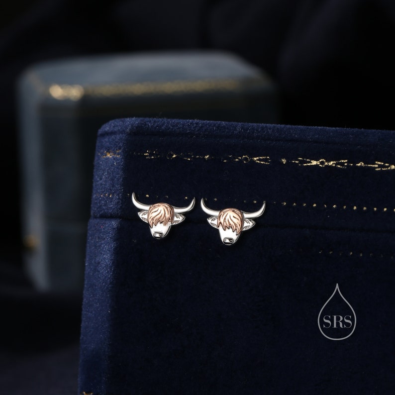 May include: A pair of silver earrings with a Highland Cow design. The earrings are on a blue velvet background.