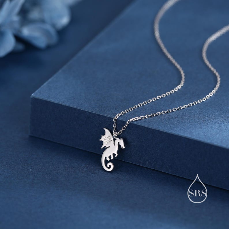 May include: A silver-colored metal necklace featuring a small dragon-shaped pendant. The dragon charm has a curled tail and wings, with the text 'SRS 925' clearly engraved on the wing. The pendant hangs from a delicate, thin silver-colored chain.