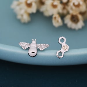 Può includere: Un paio di orecchini d'argento con un design a forma di ape e favo. L'orecchino a forma di ape ha un design dettagliato con ali e corpo. L'orecchino a forma di favo ha una piccola pietra trasparente al centro.
