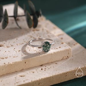 May include: A silver ring with a green moss agate stone. The ring is on a white textured surface.