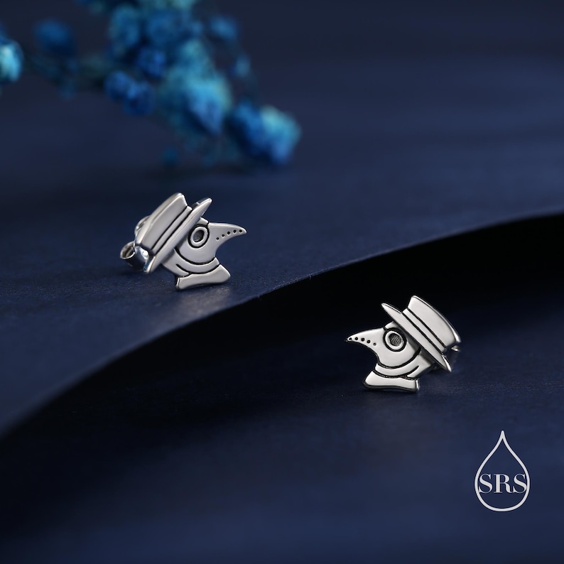 May include: Silver-toned stud earrings featuring a plague doctor design. Each earring depicts a stylized plague doctor head with a hat and beak, set against a dark blue background. The earrings are small and detailed.