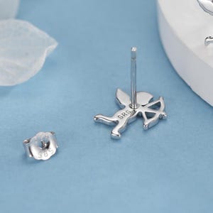 May include: Silver stud earrings with a Cupid design, complete with a bow and arrow. The earrings have a post and butterfly backing. The letters "SRS" are engraved on the Cupid's body. The earrings are on a light blue surface.