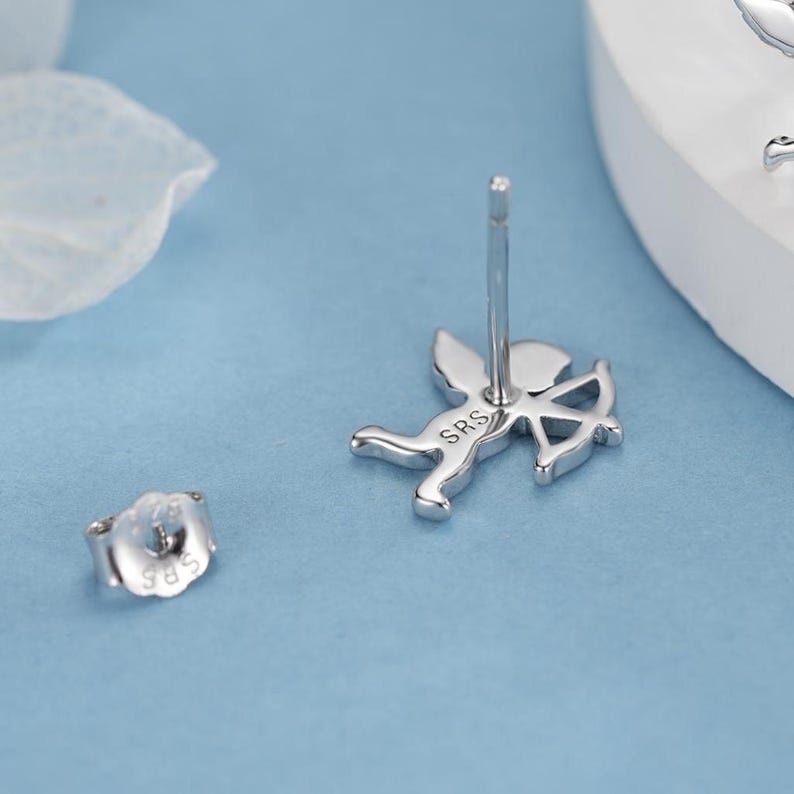 May include: Silver stud earrings with a Cupid design, complete with a bow and arrow. The earrings have a post and butterfly backing. The letters "SRS" are engraved on the Cupid's body. The earrings are on a light blue surface.