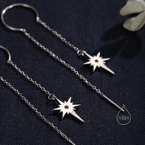 May include: Three silver dangle earrings with a star charm. The star charm has a small diamond in the center. The earrings are on a black background.