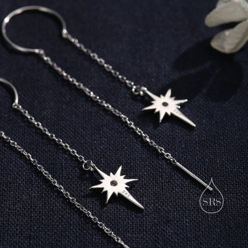 May include: Three silver dangle earrings with a star charm. The star charm has a small diamond in the center. The earrings are on a black background.