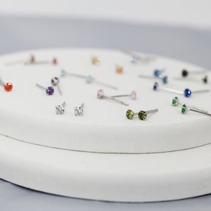 Puede incluir: Primer plano de una variedad de peque&ntilde;os pendientes de bot&oacute;n con piedras preciosas de diferentes colores. Los pendientes se muestran sobre una superficie blanca.