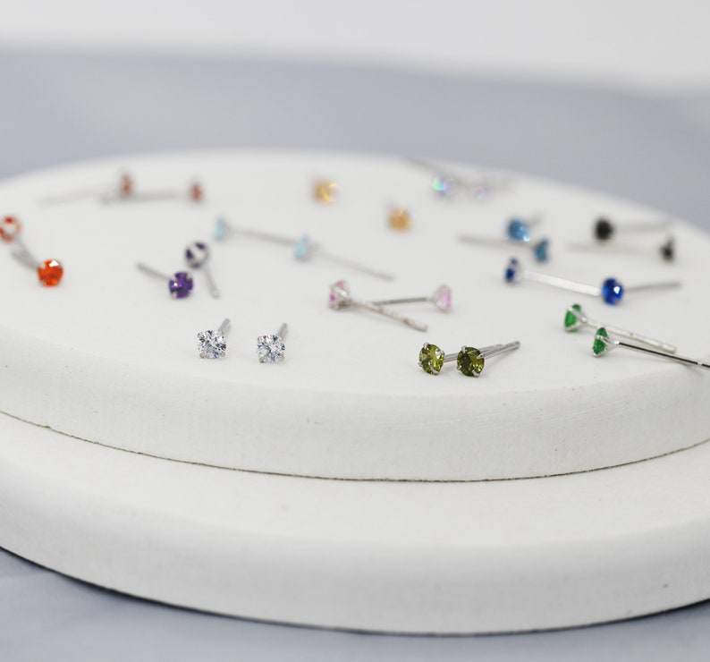 Puede incluir: Primer plano de una variedad de peque&ntilde;os pendientes de bot&oacute;n con piedras preciosas de diferentes colores. Los pendientes se muestran sobre una superficie blanca.