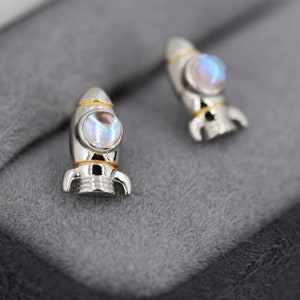 May include: A pair of silver rocket ship earrings with iridescent moonstone accents. The rockets have gold trim and are set on a gray background.