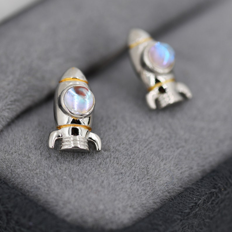 May include: A pair of silver rocket ship earrings with iridescent moonstone accents. The rockets have gold trim and are set on a gray background.