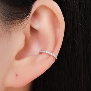 Può includere: Un ear cuff argentato con una fila di piccole pietre preziose trasparenti. Il polsino è curvo per adattarsi alla cartilagine dell'orecchio. L'immagine mostra il polsino indossato su un orecchio, con l'orecchio e la pelle circostanti visibili.