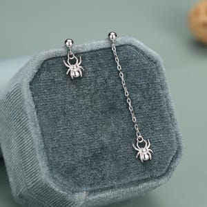 Può includere: Un paio di orecchini a lobo color argento con motivo a ragno. Un orecchino è composto da un piccolo ciondolo a forma di ragno fissato direttamente al lobo, mentre l'altro presenta una catenella lunga che termina con un ciondolo a ragno abbinato.