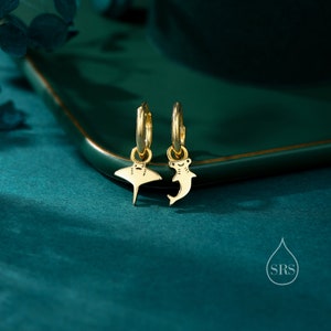 May include: A pair of gold hoop earrings with a small stingray and hammerhead shark charm hanging from each hoop. The earrings are on a green surface.
