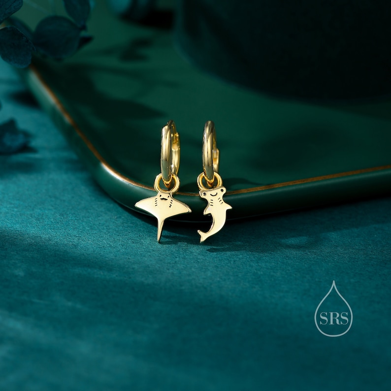 May include: A pair of gold hoop earrings with a small stingray and hammerhead shark charm hanging from each hoop. The earrings are on a green surface.