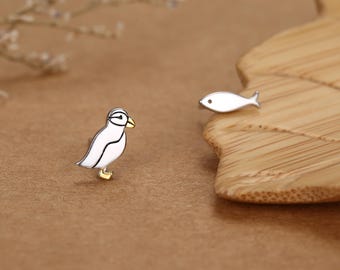 Pode incluir: Um par de brincos de tarracha com um papagaio-do-mar branco e preto com bico e patas douradas, e um pequeno brinco de tarracha em forma de peixe prateado. O papagaio-do-mar apresenta um olho preto e um contorno de asa bem definido.