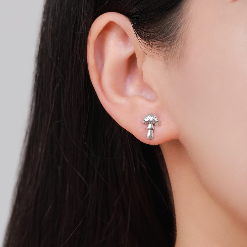 May include: A silver mushroom stud earring. The earring is a miniature mushroom design with a textured cap and a smooth stem. The earring is worn on an ear.