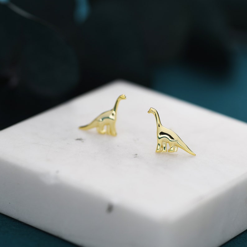 May include: A pair of gold-toned dinosaur stud earrings. The earrings are small and delicate, and they feature a simple, minimalist design.