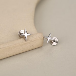 May include: A pair of silver-toned stud earrings, each shaped like a four-pointed star with elongated, tapered rays. The earrings feature a polished, reflective metallic finish and include a circular backing post clearly visible on each piece.