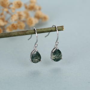 Op de afbeelding: Zilveren oorhangers met druppelvormige groene mosagaatstenen. De oorbellen hebben een gebogen haakontwerp en een kleine zilveren kraal. De achtergrond is zachtblauw met een vage tak, wat een natuurlijke uitstraling creëert.