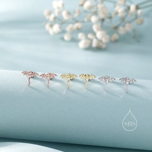 K&ouml;nnte beinhalten: Ohrstecker in Drachenform in Ros&eacute;gold, Gold und Silber. Die Ohrringe sind auf einer hellblauen Oberfl&auml;che mit einem verschwommenen Hintergrund aus wei&szlig;en Blumen dargestellt. Metallschmuck.