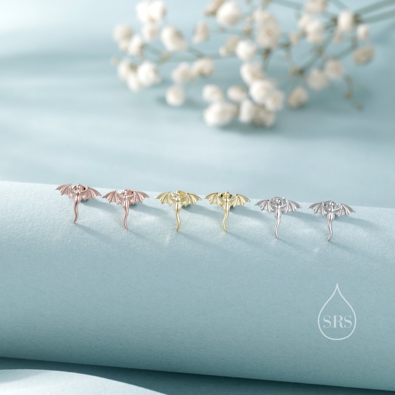 K&ouml;nnte beinhalten: Ohrstecker in Drachenform in Ros&eacute;gold, Gold und Silber. Die Ohrringe sind auf einer hellblauen Oberfl&auml;che mit einem verschwommenen Hintergrund aus wei&szlig;en Blumen dargestellt. Metallschmuck.