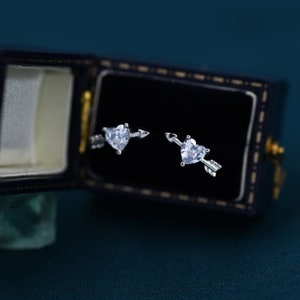 May include: A pair of silver earrings with a heart-shaped clear stone and an arrow design. The earrings are in a black velvet box.