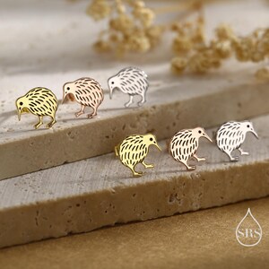 May include: A collection of kiwi bird stud earrings in gold, rose gold, and silver tones. The earrings are displayed on a textured, light-colored surface, with a soft-focus background. The kiwi bird design is detailed and stylized.