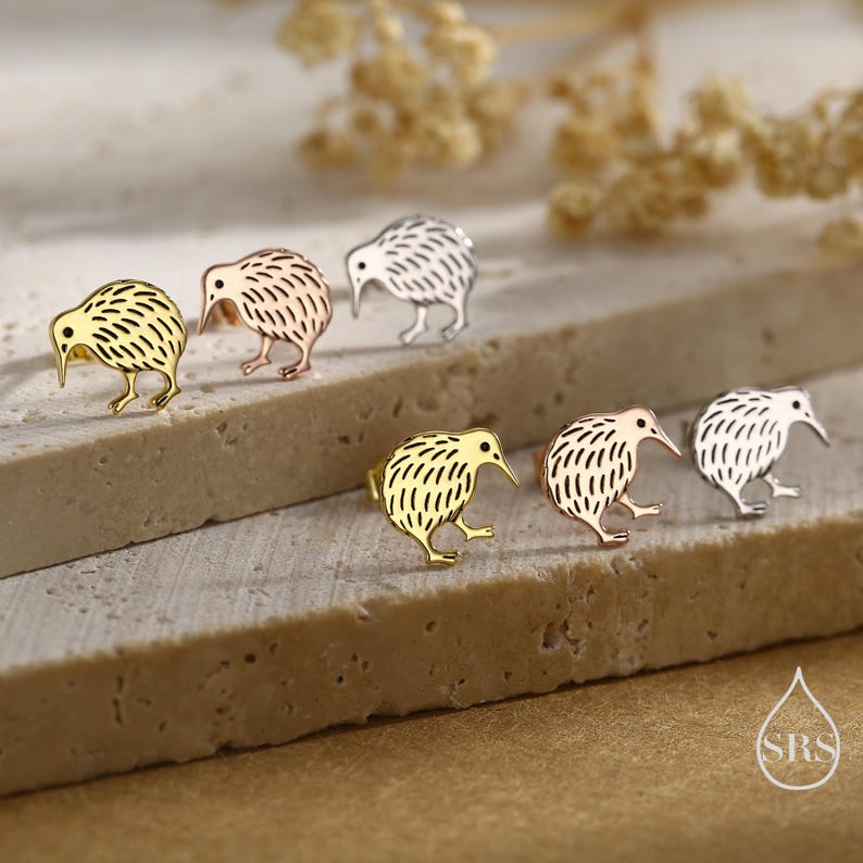 May include: A collection of kiwi bird stud earrings in gold, rose gold, and silver tones. The earrings are displayed on a textured, light-colored surface, with a soft-focus background. The kiwi bird design is detailed and stylized.