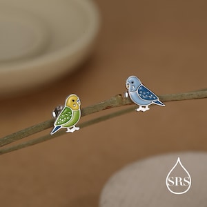 May include: A pair of silver stud earrings featuring colorful parakeet designs. One earring is yellow and green, the other is blue. The earrings are set against a blurred background, highlighting the bird-shaped jewelry.
