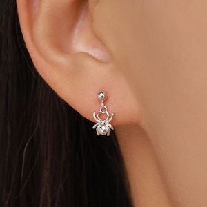 May include: A silver-toned stud earring featuring a small, dangling spider charm. The earring consists of a polished spherical stud post connected to a detailed, multi-legged spider pendant that hangs vertically from the earlobe.