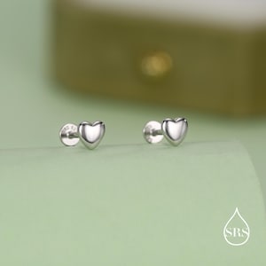 May include: A pair of silver heart-shaped stud earrings. The earrings have a polished finish and reflect light. The heart-shaped studs are mounted on posts with small backs. The background is a soft green.