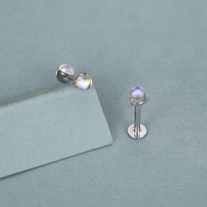 Op de afbeelding: Twee zilverkleurige labret studs met iriserende, ronde edelstenen. Eén stud staat schuin op een lichtblauwe ondergrond, de andere staat rechtop. De edelstenen tonen een mix van kleuren, waaronder geel, blauw en wit.