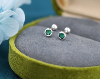 Puede incluir: Un par de pendientes de plata con pequeñas piedras preciosas verdes. Los pendientes se muestran en una caja de joyería de terciopelo verde.