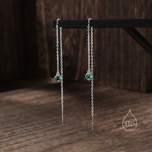 May include: A pair of silver chain earrings with green gemstone accents. The earrings hang from a black surface.