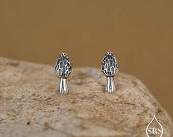 May include: A pair of silver mushroom stud earrings. The earrings feature a detailed, textured cap and a smooth stem. They are displayed on a rough, tan-coloured stone surface.
