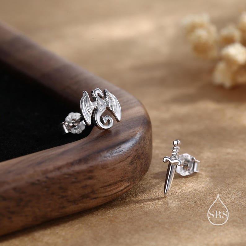 Peut inclure: Une paire de boucles d'oreilles argent&eacute;es. L'une repr&eacute;sente un dragon aux ailes d&eacute;ploy&eacute;es, l'autre une &eacute;p&eacute;e. Les boucles d'oreilles sont pr&eacute;sent&eacute;es sur une surface en bois.