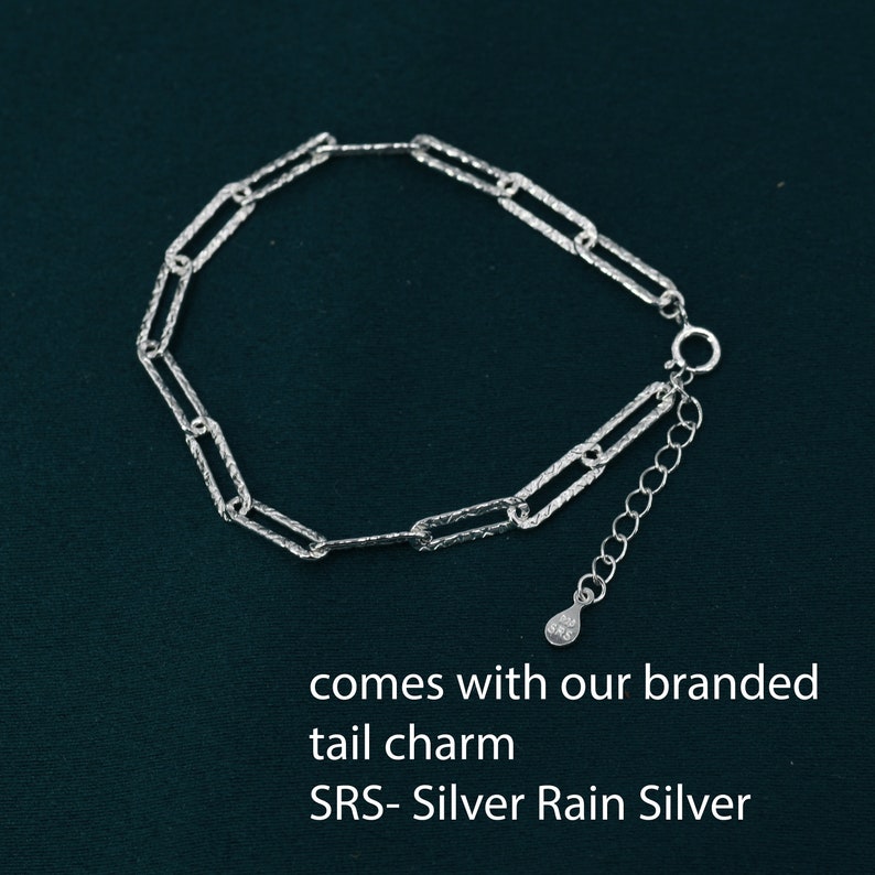 May include: A silver chain bracelet with a textured finish. The bracelet has a lobster clasp and a small silver charm with the text "SRS - Silver Rain Silver" on it.