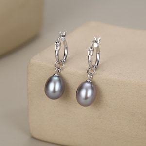 Puede incluir: Un par de pendientes de aro de plata con perlas grises colgantes en forma de lágrima. Cada pendiente consta de un aro de plata pulida con cierre de bisagra y una perla gris lisa y brillante suspendida de una pequeña anilla de plata.