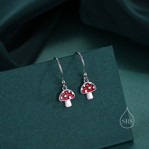 Może przedstawiać: Para srebrnych kolczyków wiszących z czerwonymi i białymi wisiorkami w kształcie grzybów. Grzyby mają białe kropki i są fantazyjnym wzorem.