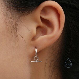 May include: Silver hoop earrings with a small bar hanging from the bottom. The earrings are shown in a person's ear.