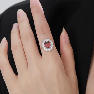 Peut inclure: Une bague en argent avec une pierre ovale rouge entourée de petites pierres claires. La bague est sertie dans un motif en forme de fleur.