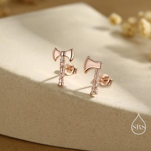 May include: A pair of rose gold axe-shaped stud earrings. The earrings have a detailed design, with the axe head and handle clearly defined. The earrings are displayed on a neutral surface, with soft lighting.