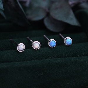 Könnte beinhalten: Vier Ohrstecker mit runden, im Cabochon-Schliff gearbeiteten Opalen, gefasst in poliertem silberfarbenem Metall. Zwei Ohrringe zeigen einen milchig-weißen Opal mit zartem Schimmer, während die anderen beiden ein leuchtend blaues Opal-Finish aufweisen.
