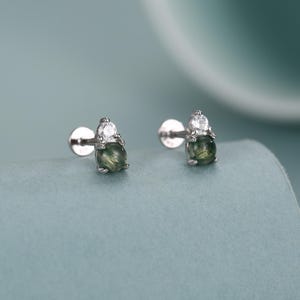 May include: A pair of stud earrings featuring a round, green gemstone base topped with a small, clear, teardrop-shaped stone. The earrings have a silver-colored post and backing, set against a soft blue-green background.
