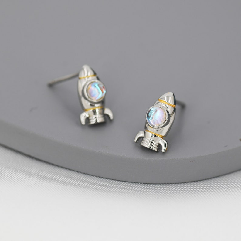 May include: A pair of silver rocket ship earrings with iridescent blue and white stones. The earrings have a gold accent around the stone.