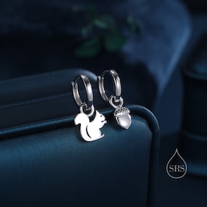 May include: A pair of silver hoop earrings with a squirrel and an acorn charm. The earrings are on a dark blue background.