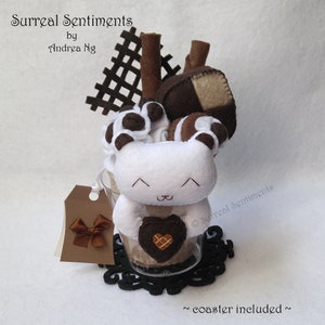 Könnte beinhalten: Ein weißes Plüschtier in Form eines Bären mit braunen Augen und einem braunen Herzen in seinen Pfoten, das in einem durchsichtigen Glasbecher mit braunen und weißen Schokoladenstücken und einem braunen Stiel obenauf sitzt. Der Becher steht auf einem schwarzen Untersetzer mit dem Text "Surreal Sentiments" und "coaster included" auf weißem Hintergrund.