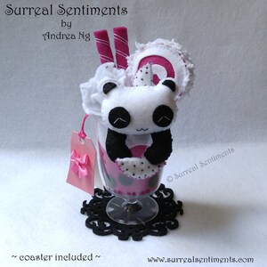 May include: A handmade panda bear plush toy in a pink and white milkshake glass with a black and white polka dot pattern. The panda is holding a pink and white lollipop. The glass is sitting on a black coaster. The text "Surreal Sentiments" is on the side of the glass.