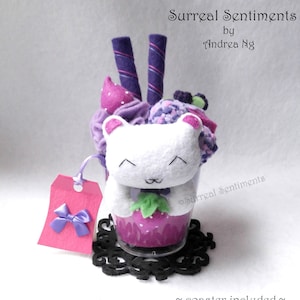 May include: A plush white bear with a pink nose and purple eyes is sitting in a clear glass cup filled with purple and pink fabric scraps. The cup is sitting on a black coaster. The text "Surreal Sentiments" is written on the cup. The text "~coaster included~" is written below the cup.